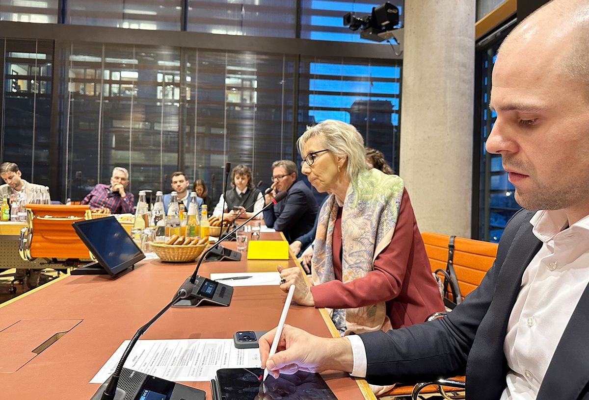 Diskussionsrunde an einem langen Tisch mit Mikrofonen; im Vordergrund eine Frau mit Brille und Schal und ein Mann, der mit einem Stift auf einem Tablet schreibt.