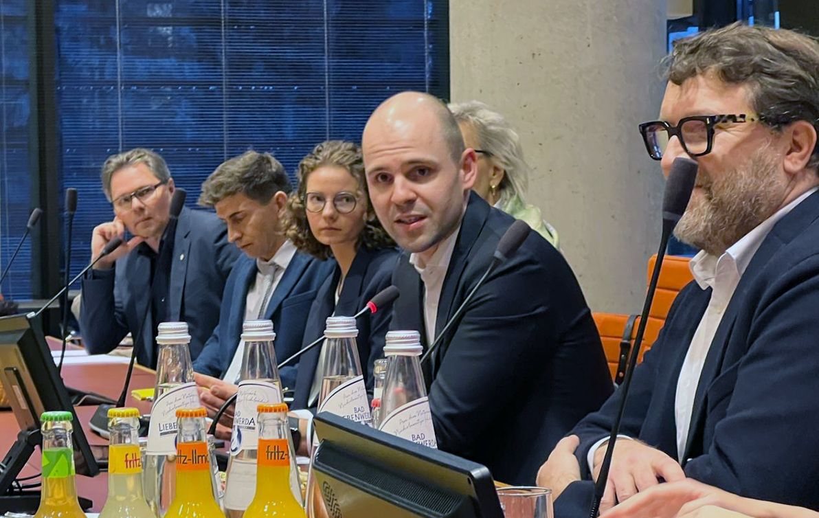 Podium des Roundtables: ein Mann spricht in ein Mikrofon, neben ihm sitzen weitere Teilnehmende an einem Tisch mit Wasserflaschen.
